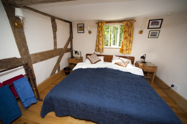 A cozy bedroom featuring exposed timber beaming, patterned yellow curtains, and a large double bed with a dark blue bedspread.