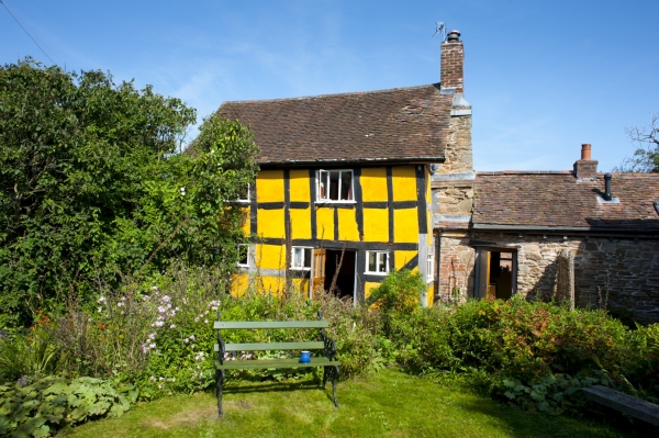 An exterior view of a picturesque yellow timber-framed cottage with a green garden bench on the lawn.
