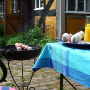 Food skewers cooking on a charcoal barbecue next to a table with a bright blue tablecloth outside the yellow timber-framed cottage.