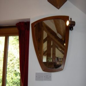 A shield-shaped wooden mirror on a white wall reflecting the exposed wooden a-frame beams and bookshelves in the bedroom.