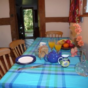 A breakfast table laid with a blue and green checkered tablecloth, blue teapot, orange juice, and a fruit bowl inside the timber-framed cottage.