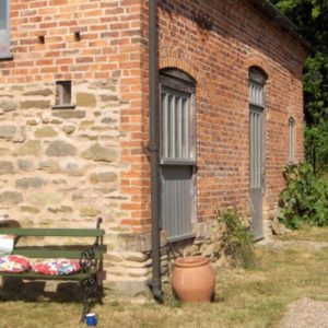 A rustic red brick outbuilding building with arched grey doors and windows next to a green garden bench.