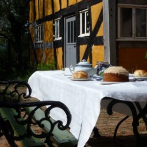 A delicious cake and hot tea set out on a table draped in a white tablecloth next to a green metal chair outside the vibrant yellow timber-framed cottage.