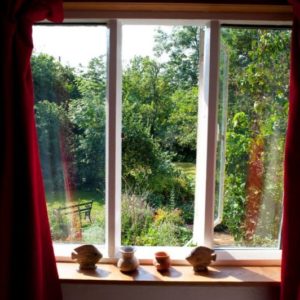 A view looking out from an open white-framed window into a lush green garden featuring trees, shrubs, and a bench.