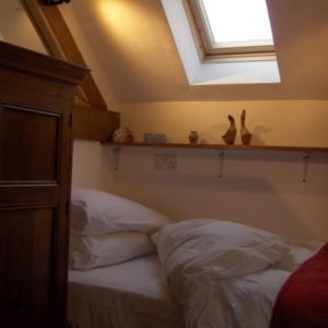 A cozy bed dressed in crisp white linens tucked under a sloping ceiling and a bright skylight, beside a wooden wardrobe.