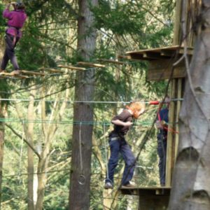 People safely navigating an elevated rope and wood obstacle course high up in the forest canopy at a Go Ape adventure park.