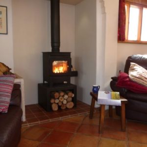 A roaring log fire glowing inside a black cast iron stove set on a tiled floor with chopped wood stacked underneath, next to a comfy armchair.