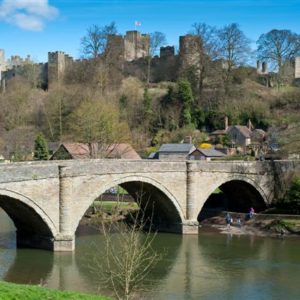 A classic stone arch bridge spanning a wide river beneath the historic Ludlow Castle situated prominently on a green hill.