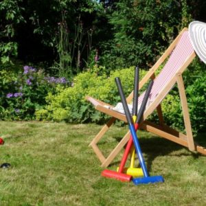 A relaxing sunny garden scene featuring a striped deckchair, a sun hat, and colorful croquet mallets resting on the grass.
