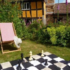 A giant outdoor chess set laid out on a checkered mat on the lawn beside a striped deckchair and the historic yellow cottage.