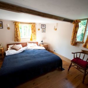 A spacious, bright bedroom with a large comfortable bed, solid oak floorboards, a dark red armchair, and exposed ceiling beams.