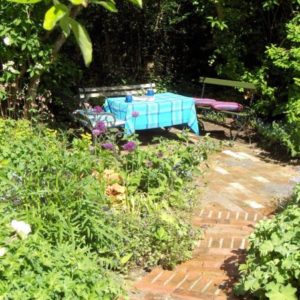 A rustic brick pathway winding through a lush green garden towards a relaxing seating area with benches and a table draped in a blue checked cloth.