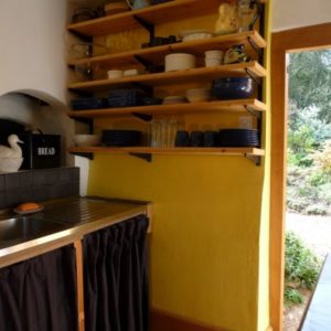 A kitchen interior featuring a vibrant yellow wall, wooden open shelves stocked with dishes, and a traditional stone bread oven opening.