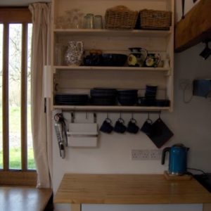 A neat kitchen area featuring a wooden butcher block table, stacked dark blue mugs, and wooden slatted wall shelves efficiently stocked with crockery.