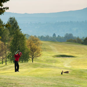 A golfer in a bright red shirt swinging a club on a verdant green golf course, backed by trees and a scenic rolling landscape fading into the distance.