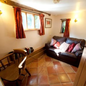 A comfortable bright living space showing a brown leather sofa, rustic red tiled flooring, and a wooden armchair beside the window.
