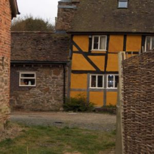 A view of the distinctive yellow timber-framed cottage and a mossy stone outbuilding, partially framed by a woven wooden hurdle fence.